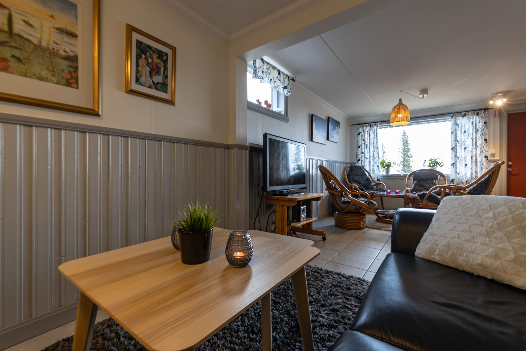 Hyggelig stue med sort sofa, sofabord i træ, TV og kurvemøbler. Vægge prydet med indrammet kunst. Delvis fortrukne gardiner, der slipper naturligt lys ind. En plante og et lys på bordet tilføjer et strejf af varme, der minder om rolige aftener i Westendorf efter et skiferieeventyr.