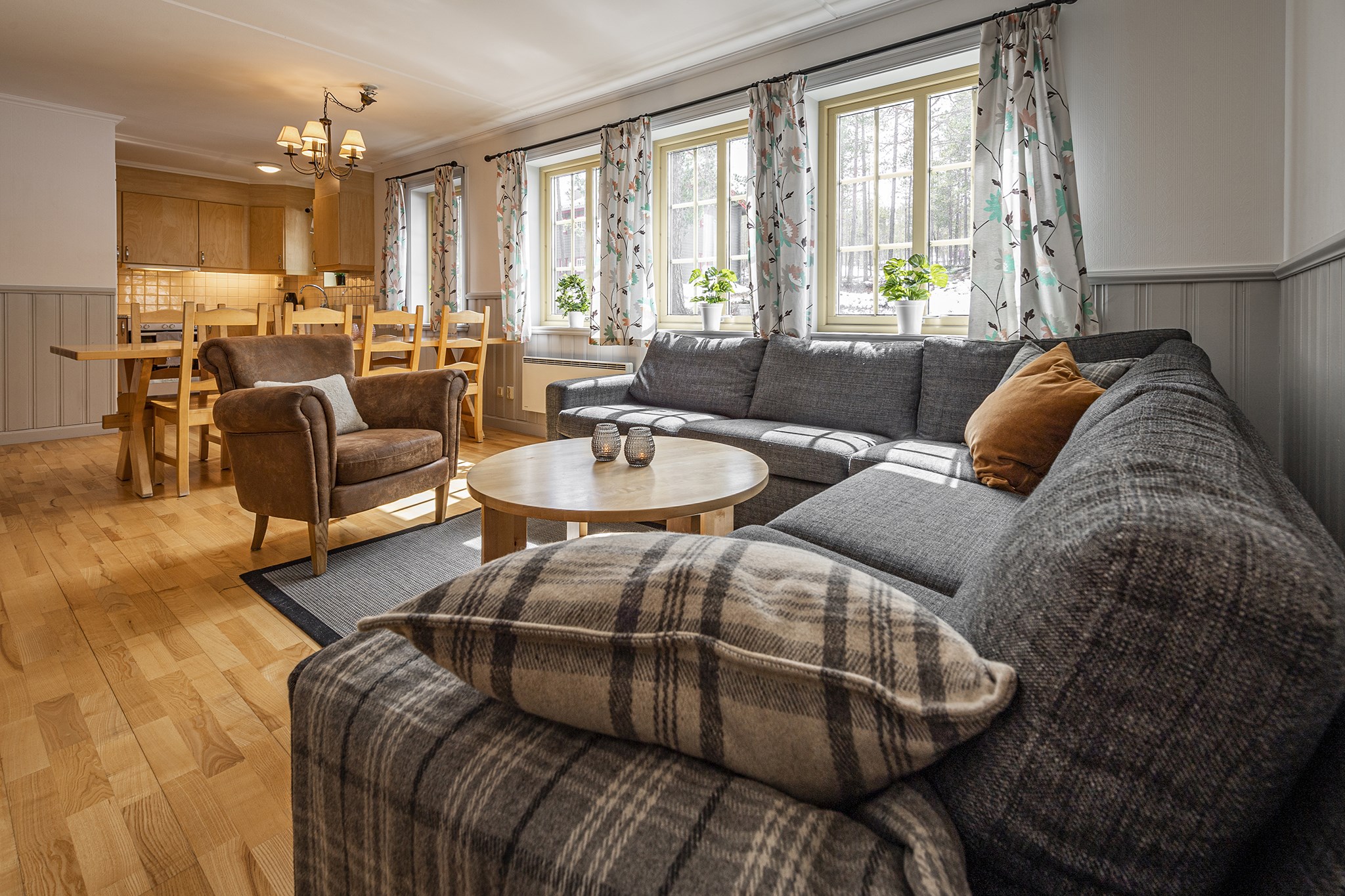Et hyggeligt stueinteriør, der minder om et sommerferiested i Wagrain, med en stor grå sektionssofa, plaidpuder og et rundt sofabord i træ. To brune lænestole vender ud mod sofaen. Et spisebord og stole af træ er i baggrunden, nær store vinduer med blomstergardiner.