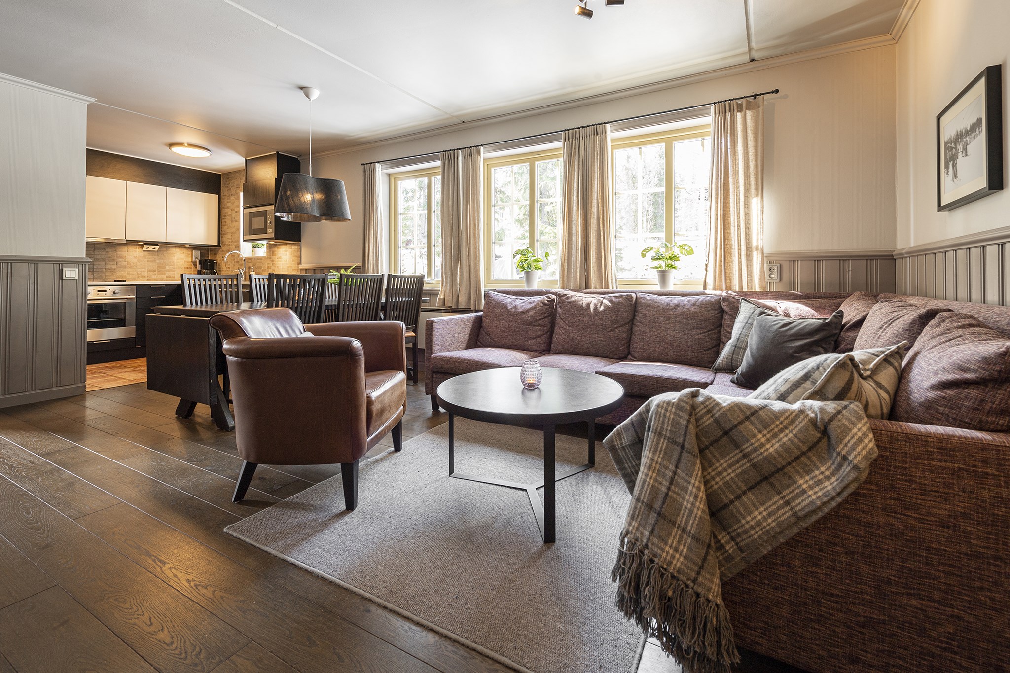 En hyggelig stue, der minder om et Westendorf-retræte, har en stor brun sektionssofa, en brun lænestol og et rundt sofabord. Et plaidkast ligger på sofaen. Et spisebord med stole er tæt på vinduet med beige gardiner, der fører til et indbydende køkken i ryggen.
