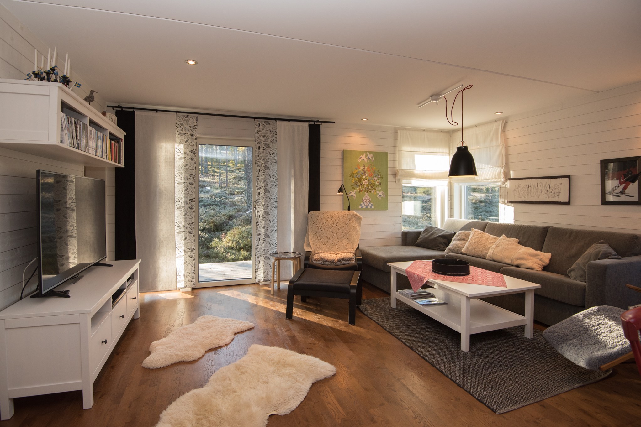 En hyggelig stue med moderne stil har en stor grå sektionssofa, et tv på en hvid stander, en lænestol og et sofabord med en rødternet løber. Inspireret af Westendorf skirejsers charme pryder fåreskindstæpper trægulvet, mens store vinduer slipper naturligt lys ind.