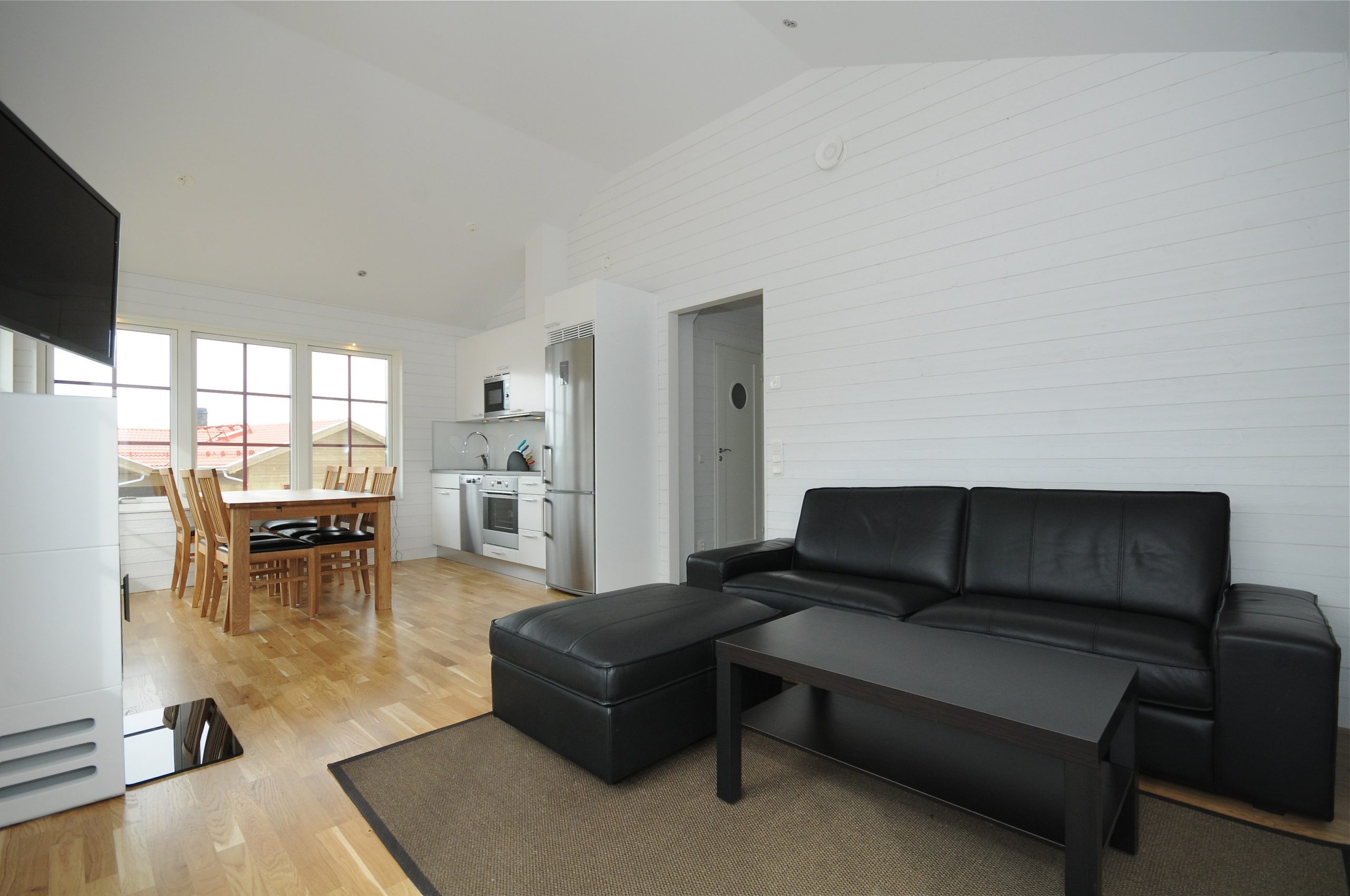 Et moderne opholds- og spiseområde med hvide vægge og loft til kip, perfekt efter en dag på pisterne i Saalbach. Den har en sort lædersofa, skammel og sofabord i træ. Et spisebord ved store vinduer tilbyder fredfyldte udsigter med et køkken i baggrunden.