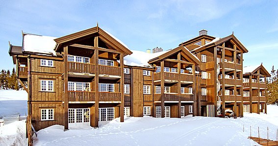 En stor træbygning i chalet-stil med flere altaner og snedækkede tage står majestætisk og minder om dem, du ville finde på skirejser i Saalbach. Strukturen er omgivet af et snelandskab under en klar blå himmel.