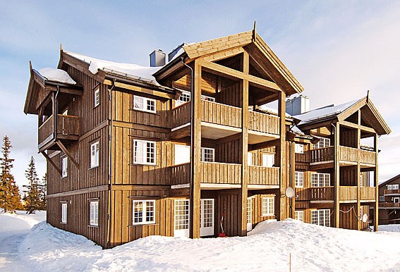 En stor træhytte med flere altaner, dækket af sne, under en klar blå himmel. Bygningen er perfekt til en skiferie og har et rustikt design med geometriske former og vinklede tage, omgivet af sne og stedsegrønne træer.