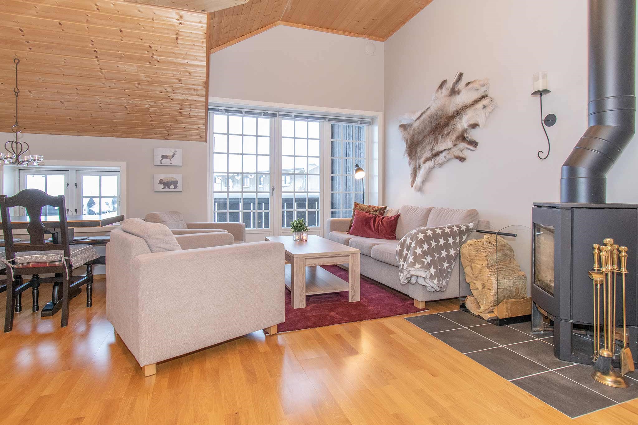 En hyggelig stue, der minder om et Saalbach-retræte, med trægulve og loft, kan prale af en brændeovn, to beige sofaer, et rødt tæppe og en spiseplads med sorte stole. Store vinduer slipper naturligt lys ind, suppleret med vægdekorationer med pels og dyreprint.