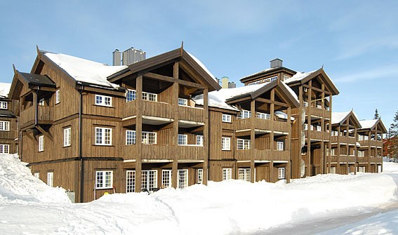 En bygning i lodge-stil i træ står midt i et snedækket landskab under en klar blå himmel, der minder om de charmerende udsigter i Westendorf. Strukturen kan prale af flere altaner og store vinduer, omgivet af dynger af sne, hvilket skaber en hyggelig vinterscene perfekt for skirejseentusiaster.