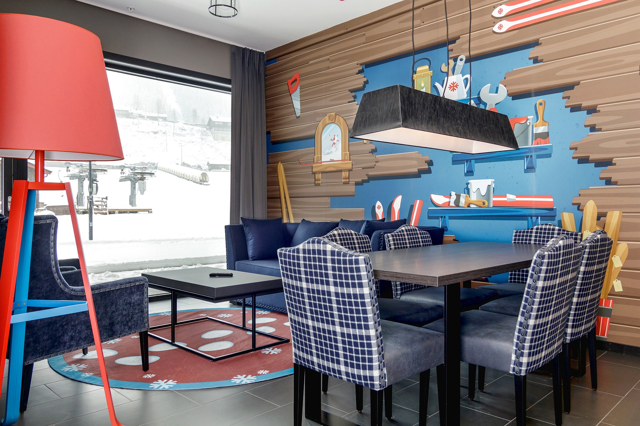 En hyggelig, moderne stue med et stort vindue, der afslører et snedækket skiferielandskab. Rummet byder på et spisebord med ternede stole, en blå sofa og en rød lampe. En væg viser legende indretning med værktøjstema, mens gulvet er prydet med et rundt tæppe, perfekt til at drømme om Wagrain-eventyr.