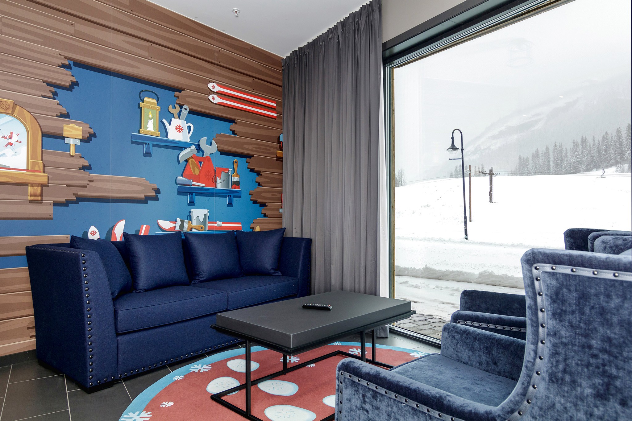En hyggelig lounge med en mørkeblå sofa og to matchende lænestole omkring et sort sofabord på et mønstret tæppe sætter scenen i dette Wagrain-inspirerede tilflugtssted. Væggen viser farverige tegnefilm af vintergenstande, mens et stort vindue afslører et snelandskab udenfor.