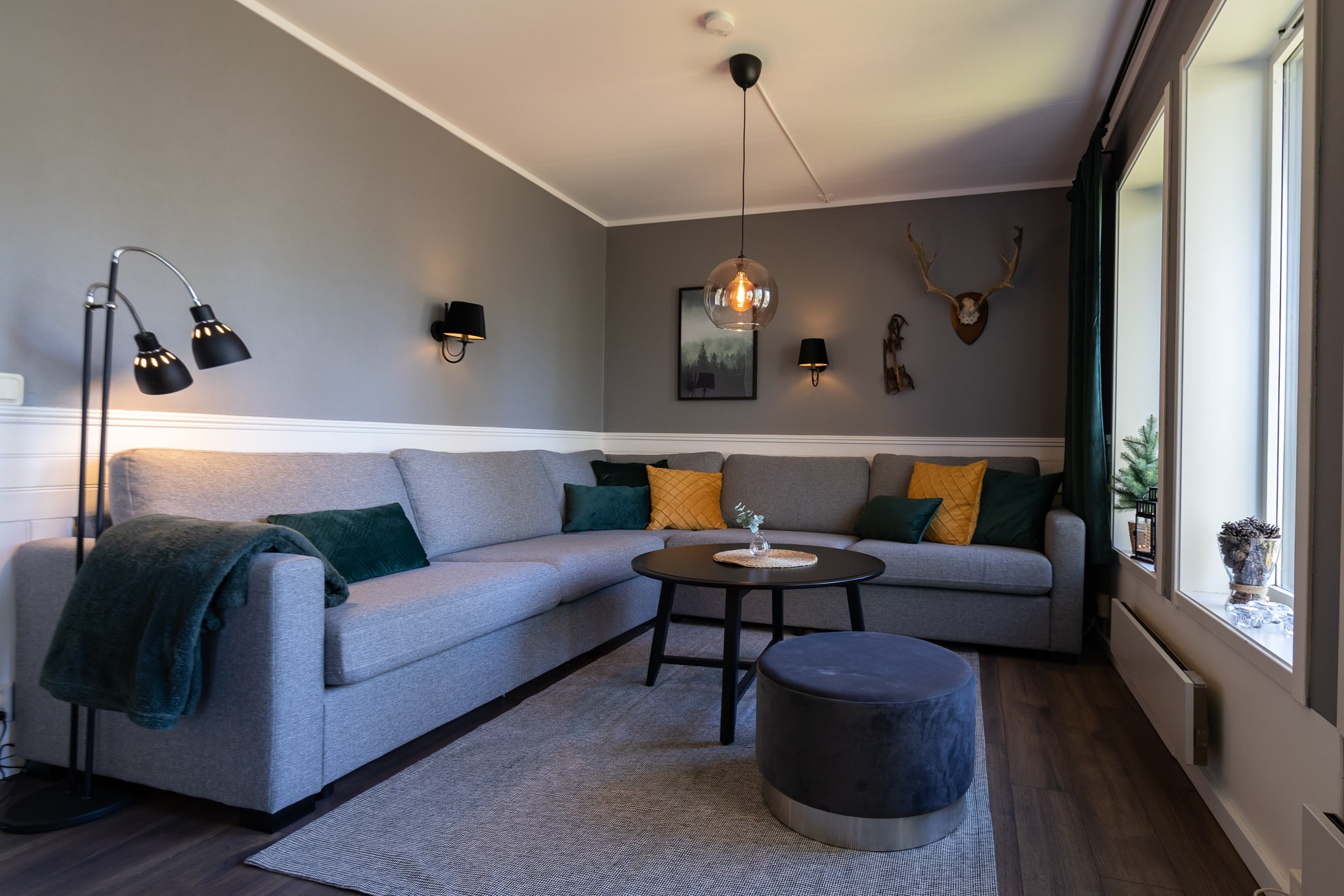 En moderne stue med en grå hjørnesofa, sort rundt sofabord og en mørkegrå skammel. Væggene er grå med monteret lys og vægdekoration, herunder et kronhjort, der minder om Wagrains alpine charme. Rummet er godt oplyst med naturligt lys fra store vinduespartier.