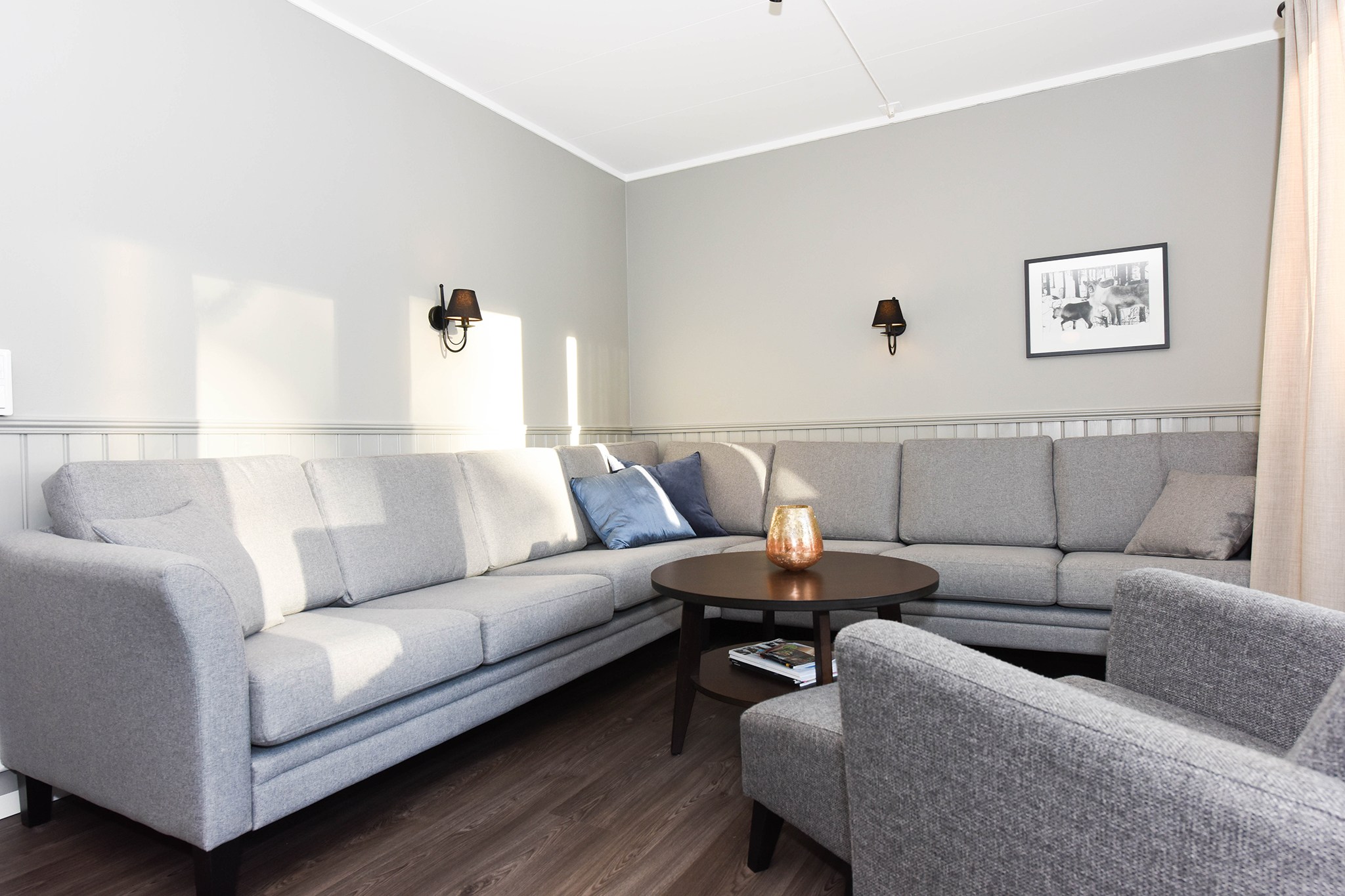 En moderne stue med en U-formet grå sofa, en grå lænestol og et rundt sofabord fremkalder den hyggelige charme af en Westendorf-hytte. Et indrammet sort-hvidt foto pryder væggen, mens to vægmonterede lamper tilbyder subtil belysning. Et gardin dækker delvist et vindue. Gulvet er af træ.