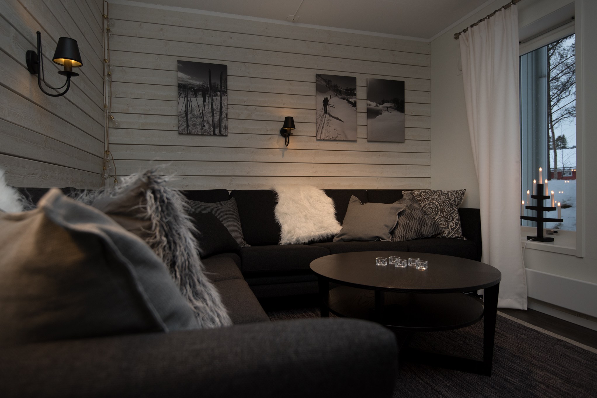 En hyggelig stue med mørk sektionssofa og forskellige teksturerede puder fremkalder varmen fra et skiferie-retræte. Sort-hvide fotos hænger på vægge med træpaneler, der minder om Westendorfs charme. Et rundt sofabord og lysestager i vindueskarmen fuldender den indbydende stemning.