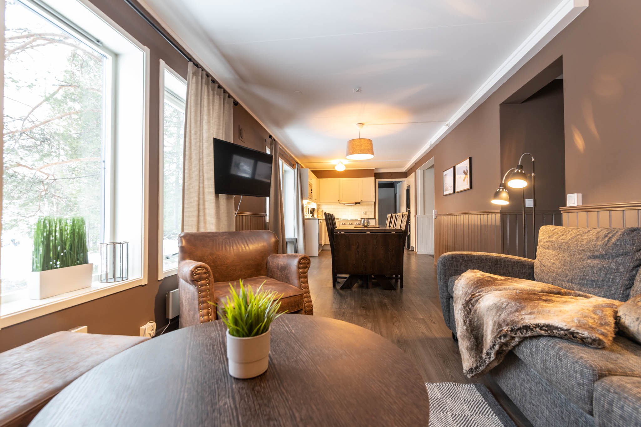 En hyggelig stue med moderne indretning, perfekt til at drømme om din næste skiferie. Den har en brun læderlænestol, en grå sofa med pels og store vinduer, der inviterer til naturligt lys. Det åbne layout fører til et køkken med moderne indretning, ideel til planlægning af skieventyr.