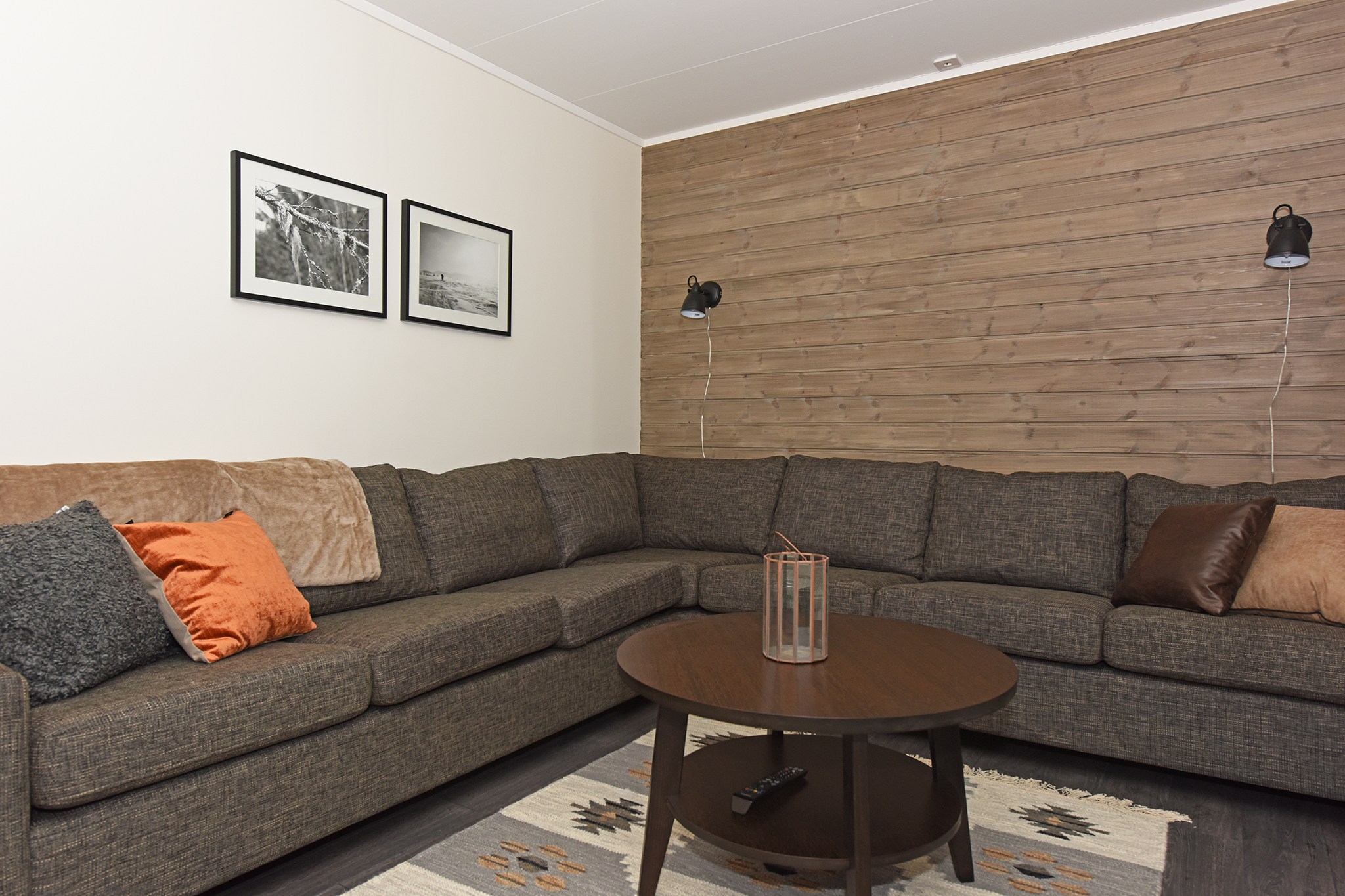 En hyggelig stue i Westendorf har en stor L-formet brun sofa prydet med orange og grå puder. Et rundt sofabord i træ sidder i midten, omgivet af vægge af træpaneler med indrammede sort-hvide fotos og vægmonterede lamper, der fremkalder en sommerferie i Østrig.