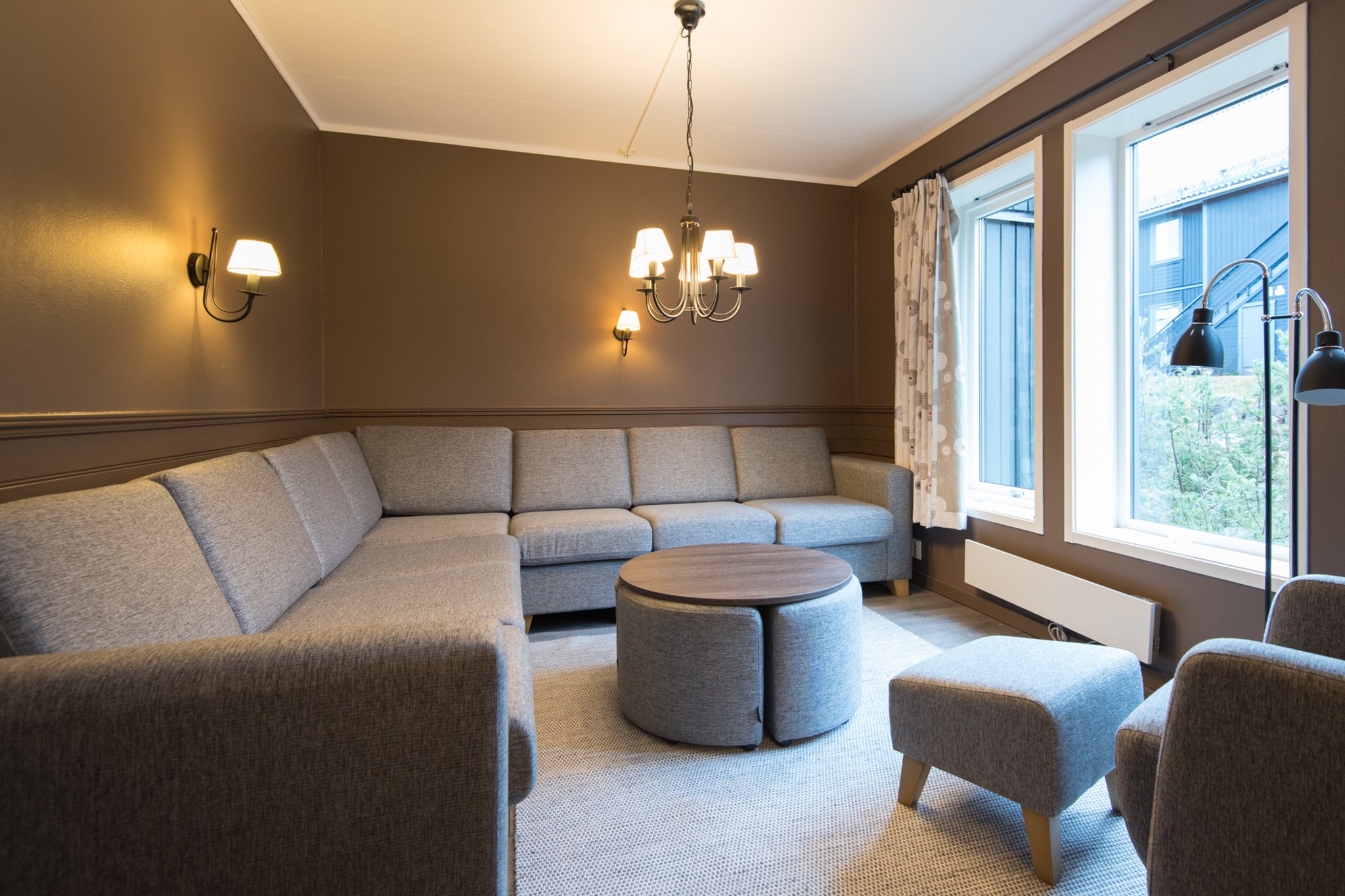 En hyggelig stue med brune vægge fanger charmen ved Wagrain, med en stor, grå L-formet sofa. Et rundt sofabord og matchende fodstøtte sidder på et beige tæppe. Elegante væglamper og en lysekrone giver blød belysning, mens høje vinduer giver udsigt udenfor.