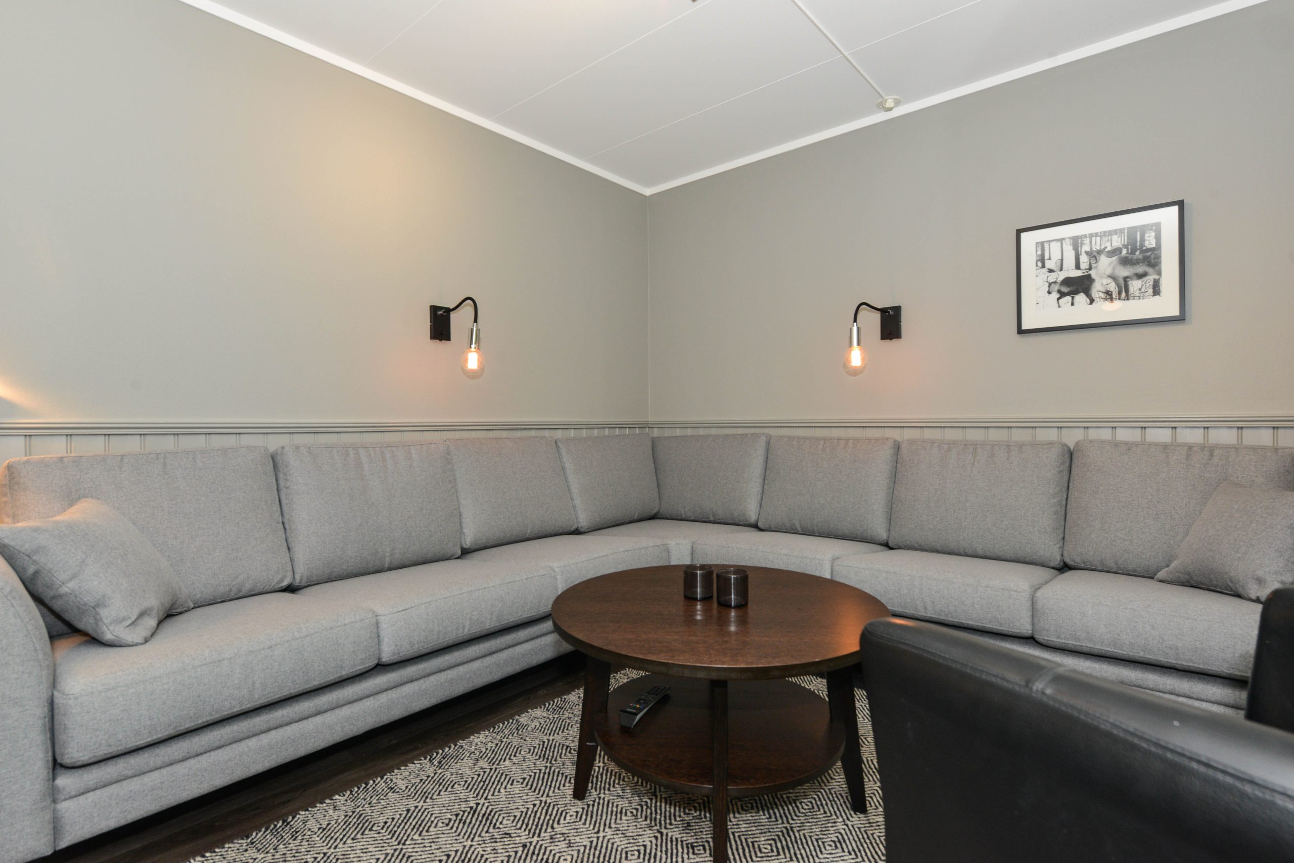 En moderne stue i hjertet af Wagrain har en stor grå L-formet sofa, et rundt sofabord i mørkt træ og to vægmonterede lysarmaturer. Et monokromt billede pryder væggen over sofaen, mens et mønstret tæppe tilføjer tekstur til dette stilfulde alpine tilbagetog.