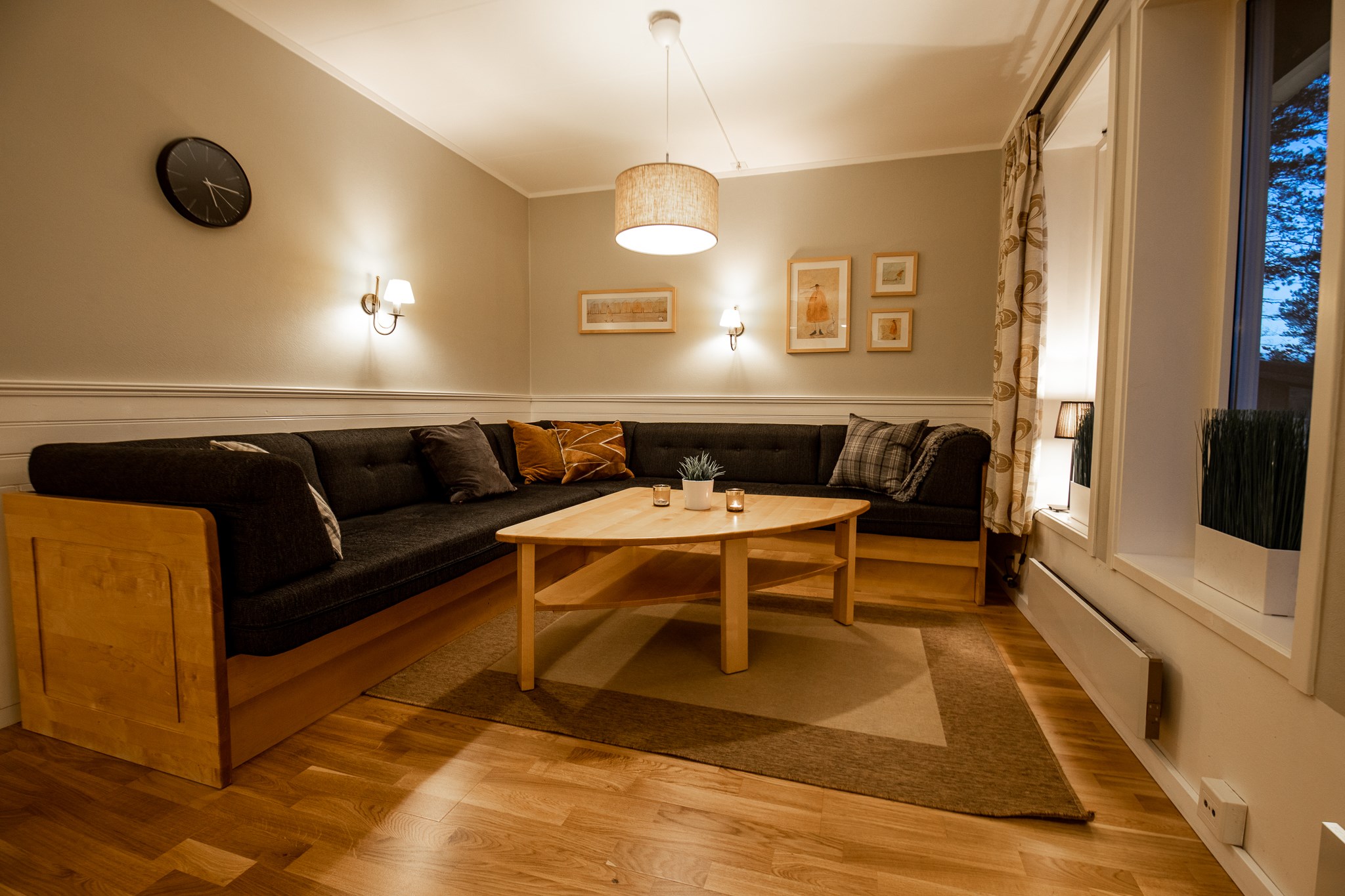 En hyggelig stue med en stor, L-formet mørk sofa, der omgiver et sofabord i træ. Væggene, der minder om en Wagrain skirejser lodge, er prydet med indrammet kunst og et ur. Varm belysning fra et loftsarmatur og væglamper oplyser rummet og fremhæver trægulvet.