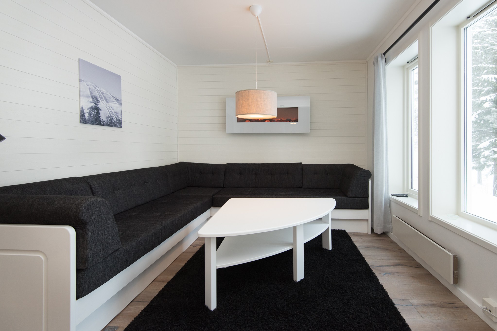En moderne stue, der minder om et skiferie-retræte, har en sort sektionssofa med hvide pynter, et hvidt trekantet sofabord på et sort tæppe og et stort vindue. Væggene er hvide, prydet med et landskabsfotografi, og fra loftet hænger en beige pendel.