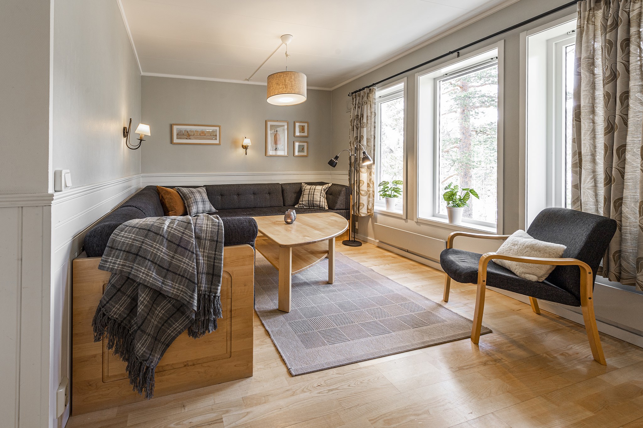 En hyggelig stue, der minder om et Wagrain-retræte, med en stor L-formet sofa, sofabord i træ og moderne lænestol. Lyst trægulv, et stort vindue med gardiner og vægkunst forstærker skirejser-stemningen. Et gråt område tæppe og hængende loftslampe fuldender dette indbydende rum.