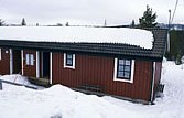 En rød hytte med snedækket tag står midt i et snelandskab i Saalbach. Mørk-indrammede vinduer pryder trævæggene, og stedsegrønne træer er synlige i baggrunden under en overskyet himmel, perfekt til dem, der drømmer om skirejser.