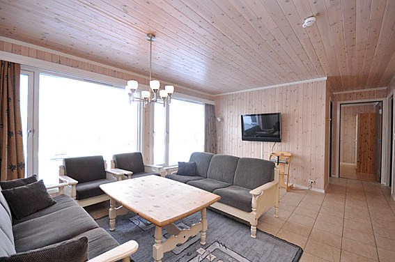 En hyggelig stue med trævægge og loft, der minder om en Saalbach skiferie, med grå polstrede sofaer omkring et lyst sofabord i træ. Et vægmonteret tv og store vinduer tillader naturligt lys at fylde rummet, perfekt til at slappe af efter en dag på pisterne.
