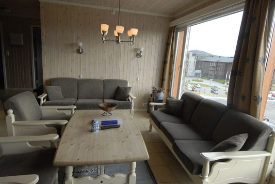 En hyggelig stue med trævægge og store vinduer fanger charmen ved et Saalbach-retræte. To grå sofaer og lænestole omkranser et sofabord i træ under en lysekrone. Vinduerne indrammer perfekt en udendørs scene med bygninger og parkerede biler, hvilket indbyder til tanker om sommerferie Østrig.