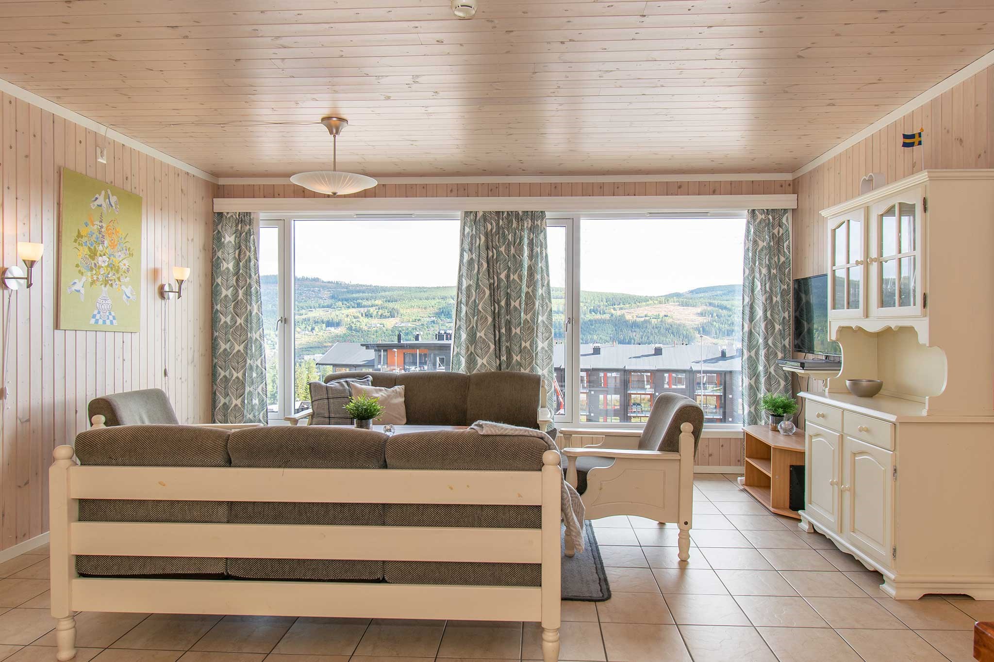 En hyggelig stue med træloft og store vinduespartier med en naturskøn udsigt over Saalbach-bakkerne. Der er en grå sofa, en hvid lænestol, et tv på et skab og et farverigt vægmaleri. Lysgardiner indrammer vinduerne, perfekt til at mindes om sommerferie Østrigs eventyr.
