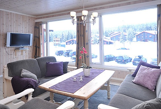 En hyggelig stue med træloft og store vinduer åbner ud til et sneklædt Saalbach-landskab. Værelset har grå sofaer med lilla hynder, et sofabord i træ med en lilla løber, en lille plante og et væghængt tv, perfekt til at koble af efter skiferieeventyr.