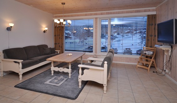 En hyggelig stue i Wagrain har en mørk sofa og lænestole omkring et sofabord i træ på et mønstret tæppe. Store vinduer afslører en sneklædt scene, perfekt til afterski-afslapning. De lyspanelede vægge forstærkes af et fladskærms-tv monteret på højre væg.