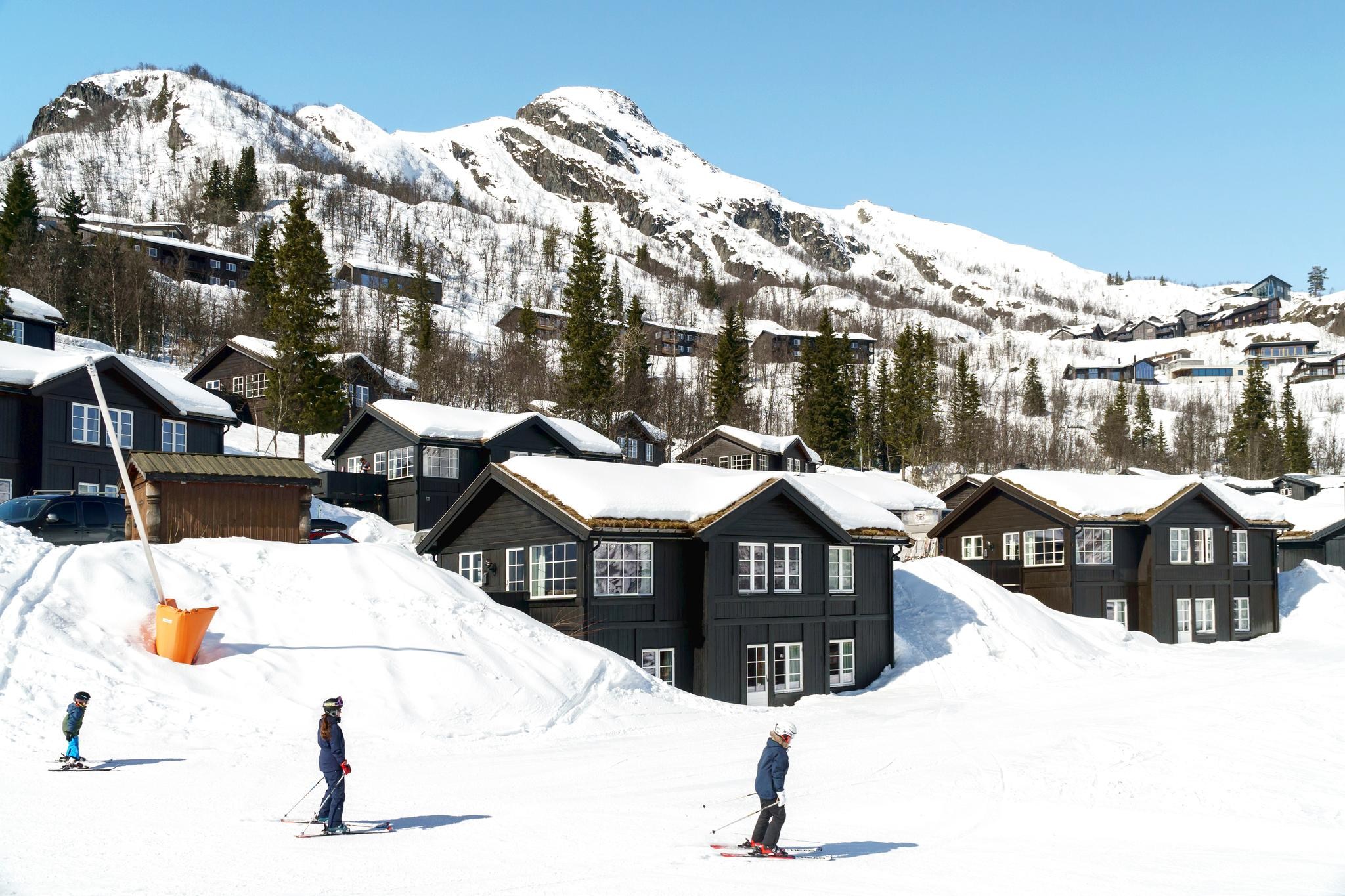 Skiløbere glider ned ad en sneklædt skråning foran mørke træhytter i Westendorf. Snedækkede bjerge og fyrretræer danner baggrunden under en klar blå himmel, hvilket gør denne skiferie uforglemmelig.