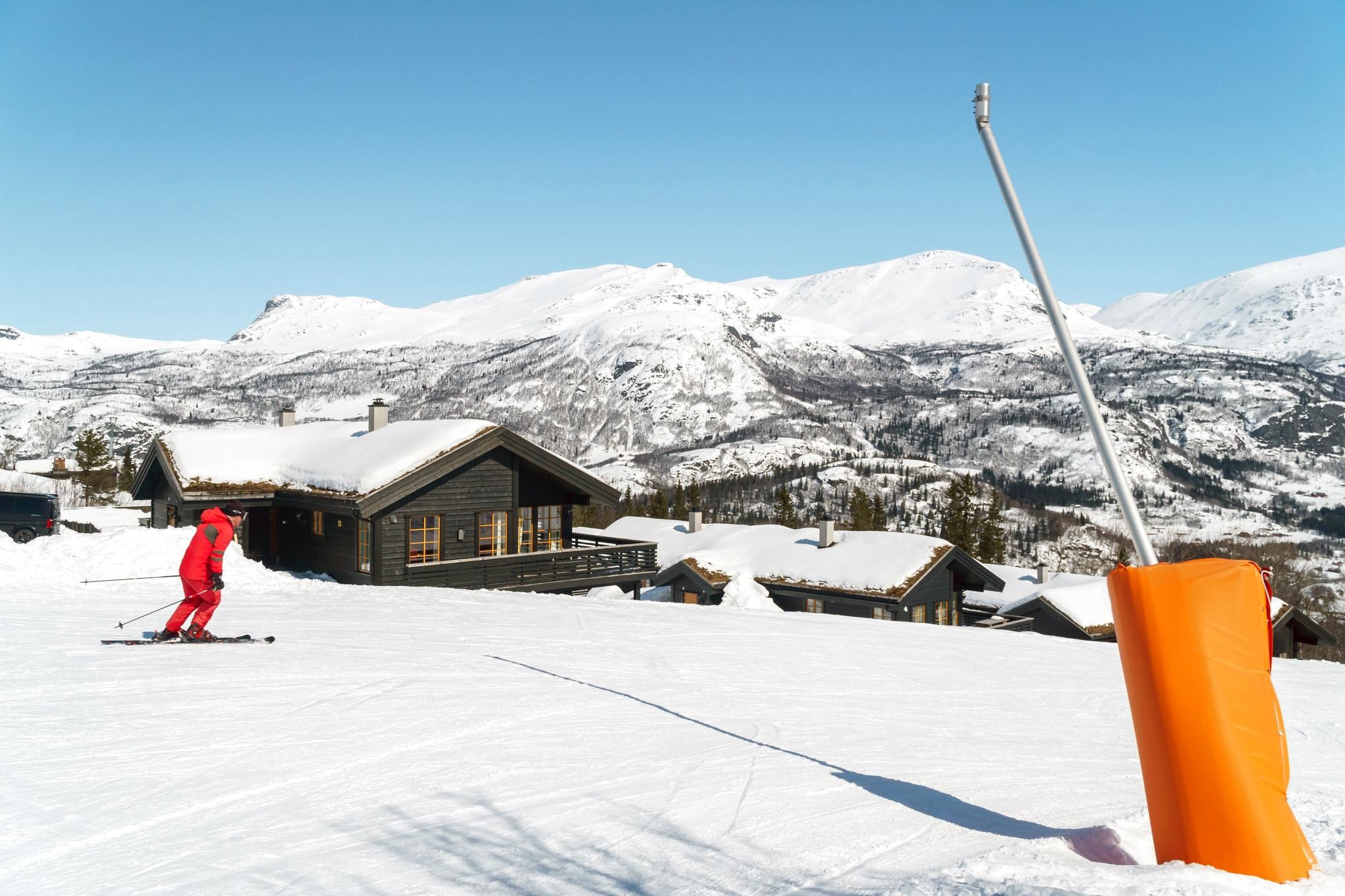En skiløber i rødt outfit går ned ad en sneklædt skråning nær træhytter og fanger essensen af en skiferie. Snedækkede bjerge og træer pryder baggrunden under en klar blå himmel, der minder om maleriske Saalbach. En orange markeringsstang står fremtrædende i forgrunden.