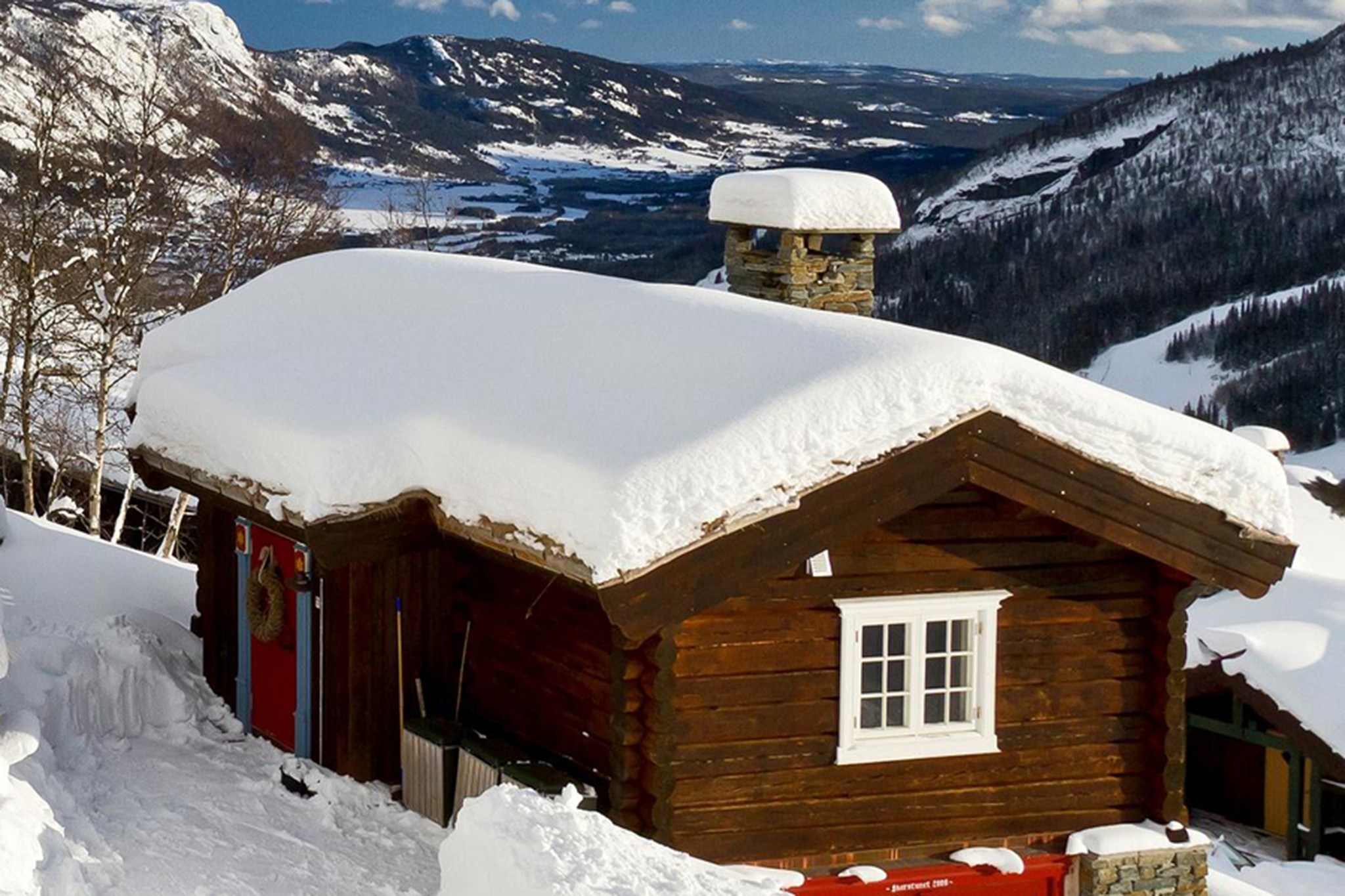 En hyggelig træhytte med snedækket tag ligger i et sneklædt bjerglandskab, perfekt til en skiferie. En stenskorsten kigger gennem sneen, mens nøgne træer og fjerne bjerge maler baggrunden under Saalbachs klare blå himmel.