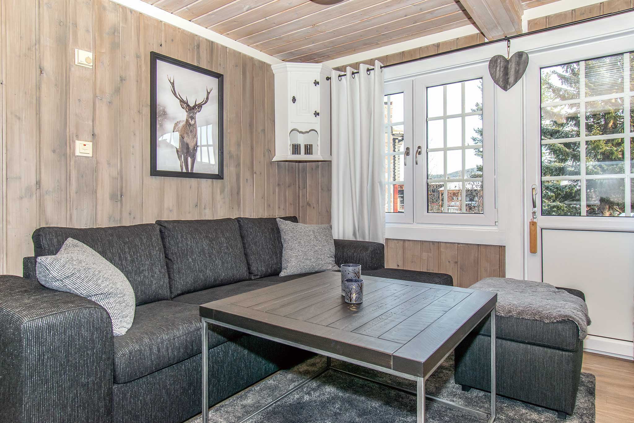 Hyggelig stue med mørkegrå sektionssofa og matchende skammel, perfekt til afterski-afslapning efter en dag i Saalbach. Et sofabord i træ sidder i midten, mens et indrammet hjortebillede hænger på væggen med træpaneler. Lysgardiner indrammer vinduet og døren, med en hjerteformet dekoration i nærheden.