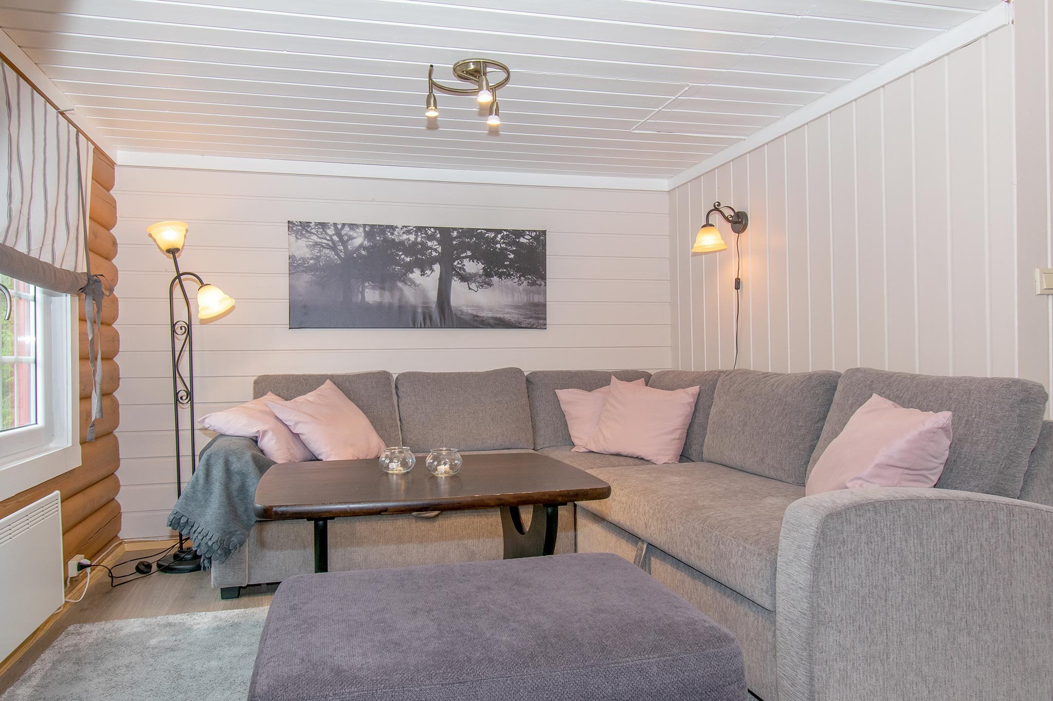 Hyggelig stue inspireret af Saalbachs alpine charme, med en grå sektionssofa og lyserøde hynder. Et sofabord af træ og en lilla skammel står i centrum med væglamper, en gulvlampe og et indrammet skovfoto på væggen. Rummet har et lyst træpanelloft, der minder om skirejser-retreater.