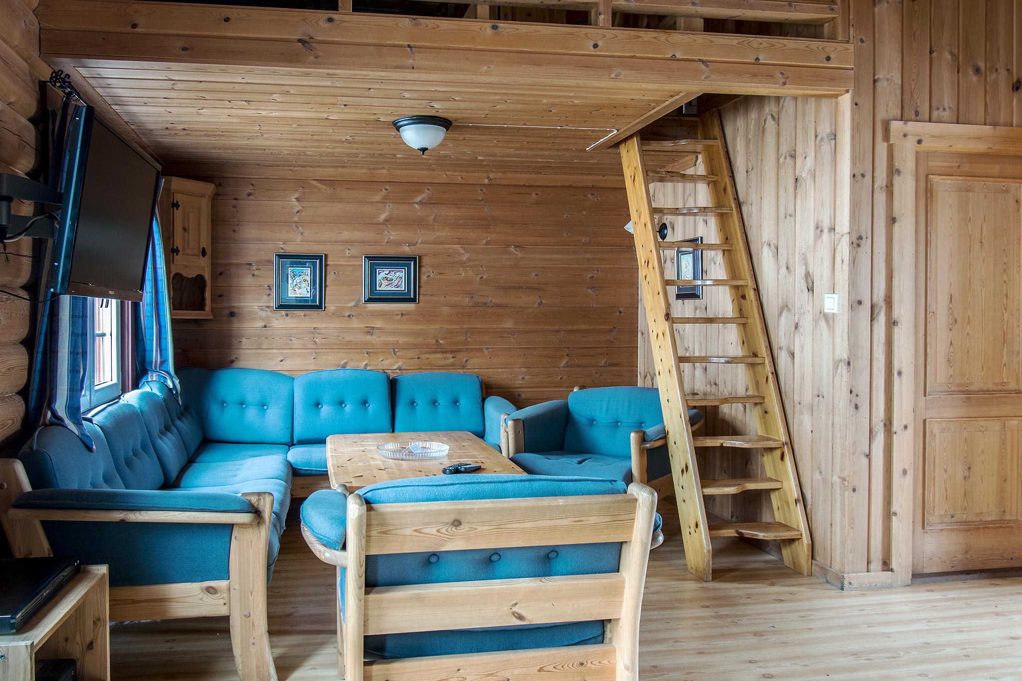 En hyggelig træstue med blågrønne polstrede møbler, herunder en sofa og stole omkring et sofabord i træ. En træstige fører til en øvre hems. Perfekt til et skiferie-retræte, værelset har en rustik hyttestemning, der minder om charmerende Saalbach- eller Wagrain-hytter.