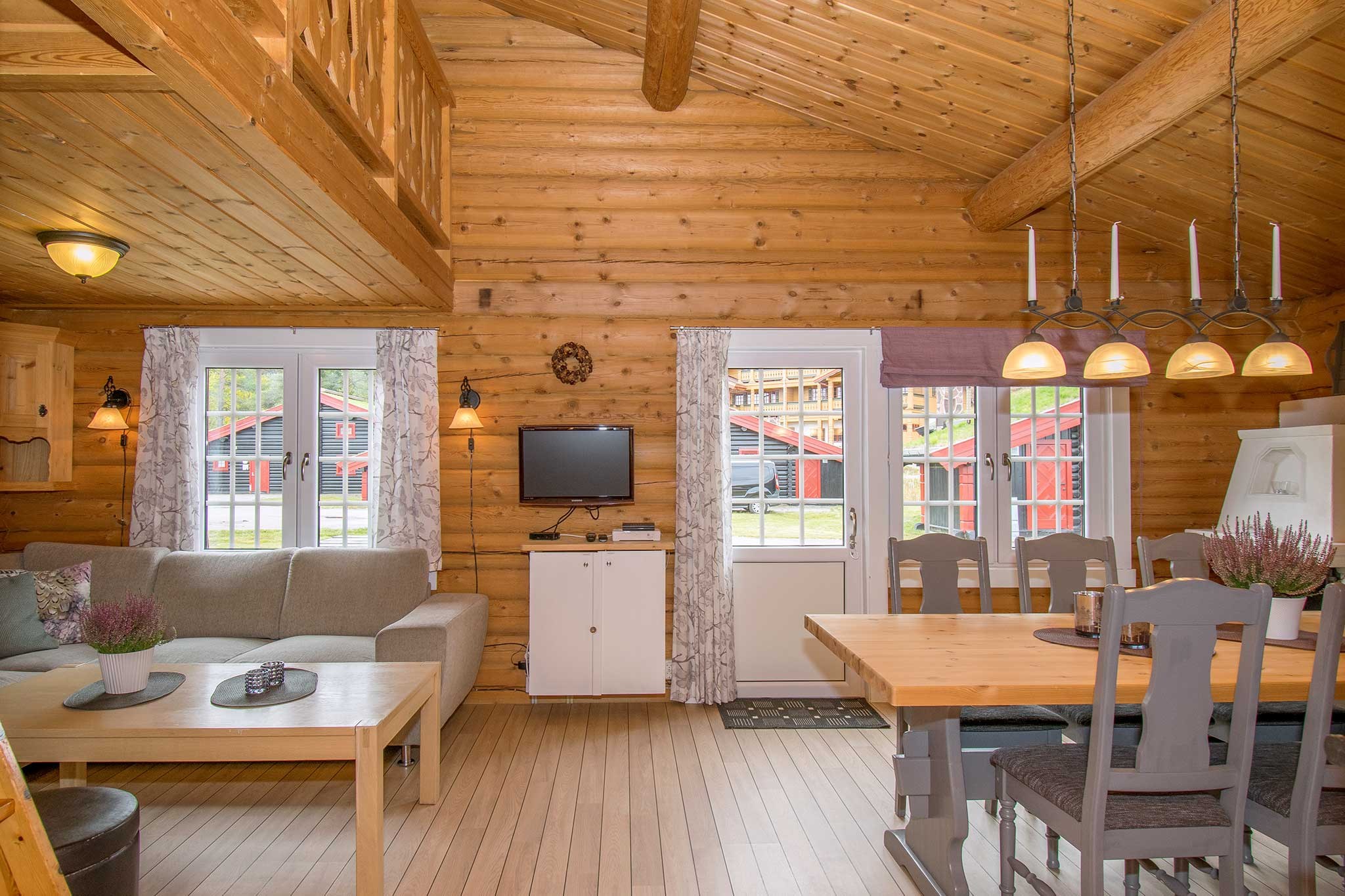 Et hyggeligt bjælkehytteinteriør med trævægge og -lofter, perfekt til en skiferieflugt. Det har et opholdsområde med sofa, et lille tv og et spisebord med stole. En dør og vinduer åbner til en udendørs udsigt, der minder om Wagrains charme. Pendellamper hænger over spisebordet.