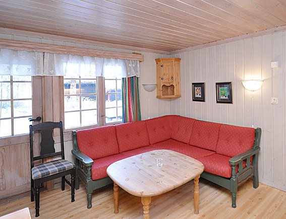 En hyggelig stue med trævægge og loft, der minder om en Westendorf skiferie. Der er en rød polstret hjørnesofa med et sofabord i træ foran. En plaid stol er ved siden af vinduet. To små indrammede kunstværker hænger på væggen, som lys filtrerer gennem vinduesgardinerne.