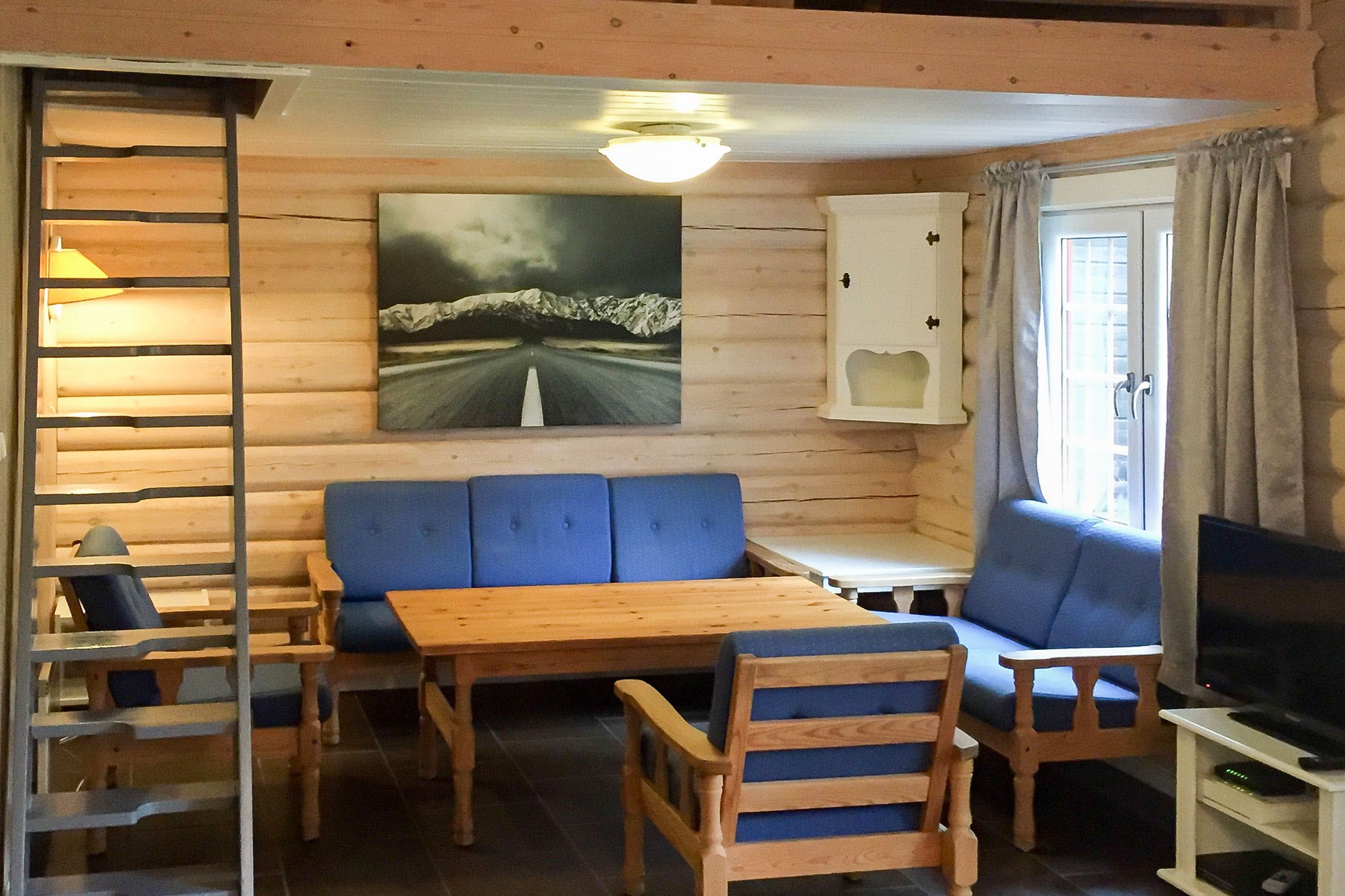 Hyggelig kabineindretning med trævægge og møbler, perfekt til en skirejser. Et opholdsområde med blå polstrede stole og en sofa omkring et træbord. Et maleri af Westendorfs vej og bjerge pryder væggen. Naturligt lys filtrerer ind gennem et vindue og oplyser stigen til hemsen.