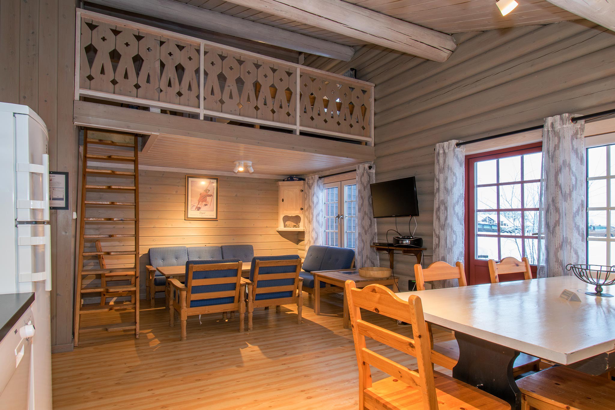 Hyggeligt træinteriør i en hytte i Westendorf med hems, perfekt til din skiferie. Har et spisebord med stole, et opholdsområde med en blå sofa og en trappe, der fører til hemsen. Store vinduer giver naturligt lys og tilbyder udsigt over det sneklædte landskab udenfor.