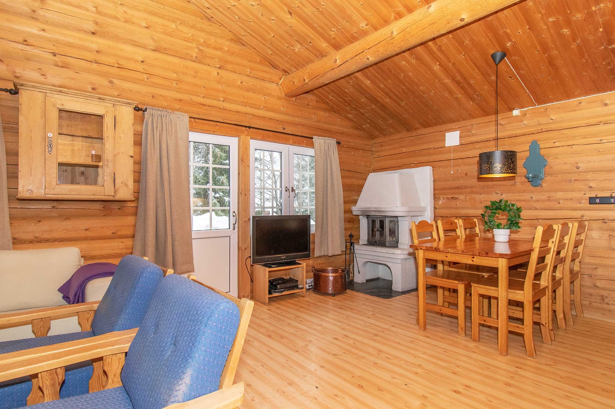 Et hyggeligt bjælkehytteinteriør med trævægge og -loft fanger charmen ved Wagrain. Et spisebord med stole står ved siden af en hvid pejs, perfekt til afslappende afterski eller sommerferie Østrig. Et tv hviler i nærheden af dobbeltdøre, der fører udenfor, mens blå polstrede stole og en grøn plante fuldender den rustikke indretning.