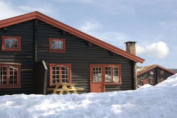 En træhytte med rød trim sidder delvist dækket af sne under en blå, let overskyet himmel. Perfekt til skirejse-entusiaster, skorstenen er synlig og et picnicbord hviler ved siden af. En anden lignende hytte titter frem i baggrunden, der minder om hyggelige ophold i Westendorf under din sommerferie Østrig.