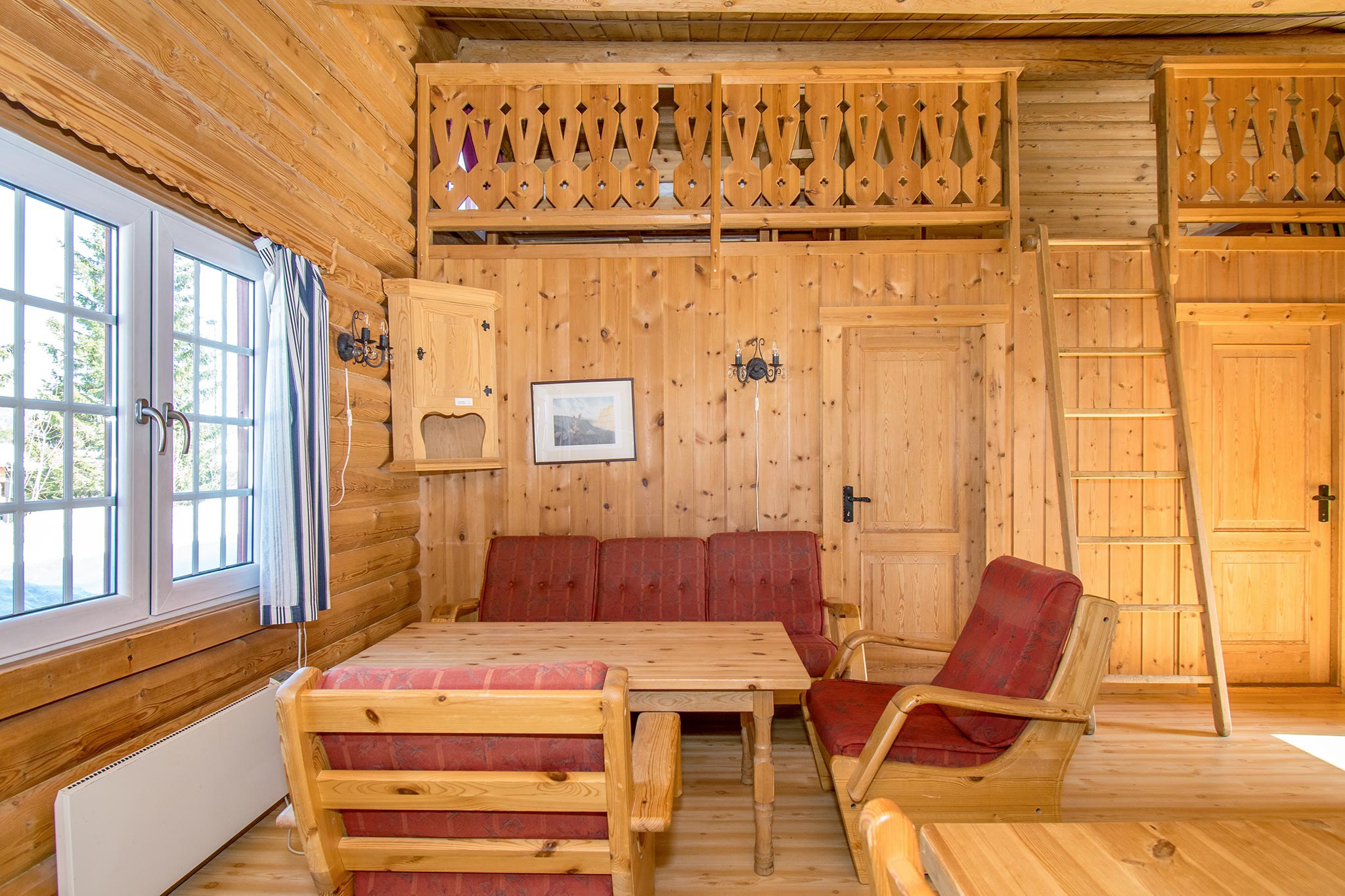 Hyggelig bjælkehytte interiør med trævægge og loft, der minder om Westendorf charme. Værelset har rødpudede træstole, et matchende bord, hems med stige og et vægmonteret billede. Naturligt lys strømmer gennem et vindue med blåstribede gardiner.