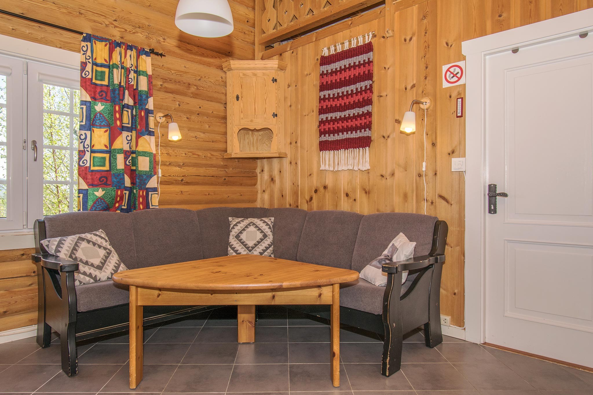 Et hyggeligt træværelse, der minder om et charmerende alpine tilbagetog, med en hjørnesofa og to mønstrede hynder. Foran sidder et trekantet træbord og vækker minder om skirejser i Saalbach. Et vævet gobelin pryder væggene, flankeret af farverige gardiner og et rygeforbud på den hvide dør.