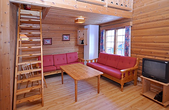 Et hyggeligt bjælkehytteinteriør, der minder om et tilbagetog i Westendorf, med trævægge og gulvbelægning. Den har røde polstrede træsofaer, et sofabord i træ og en stigelignende trappe, der fører til et øvre område. Et lille tv står på et stativ i hjørnet, og vinduer giver naturligt lys.