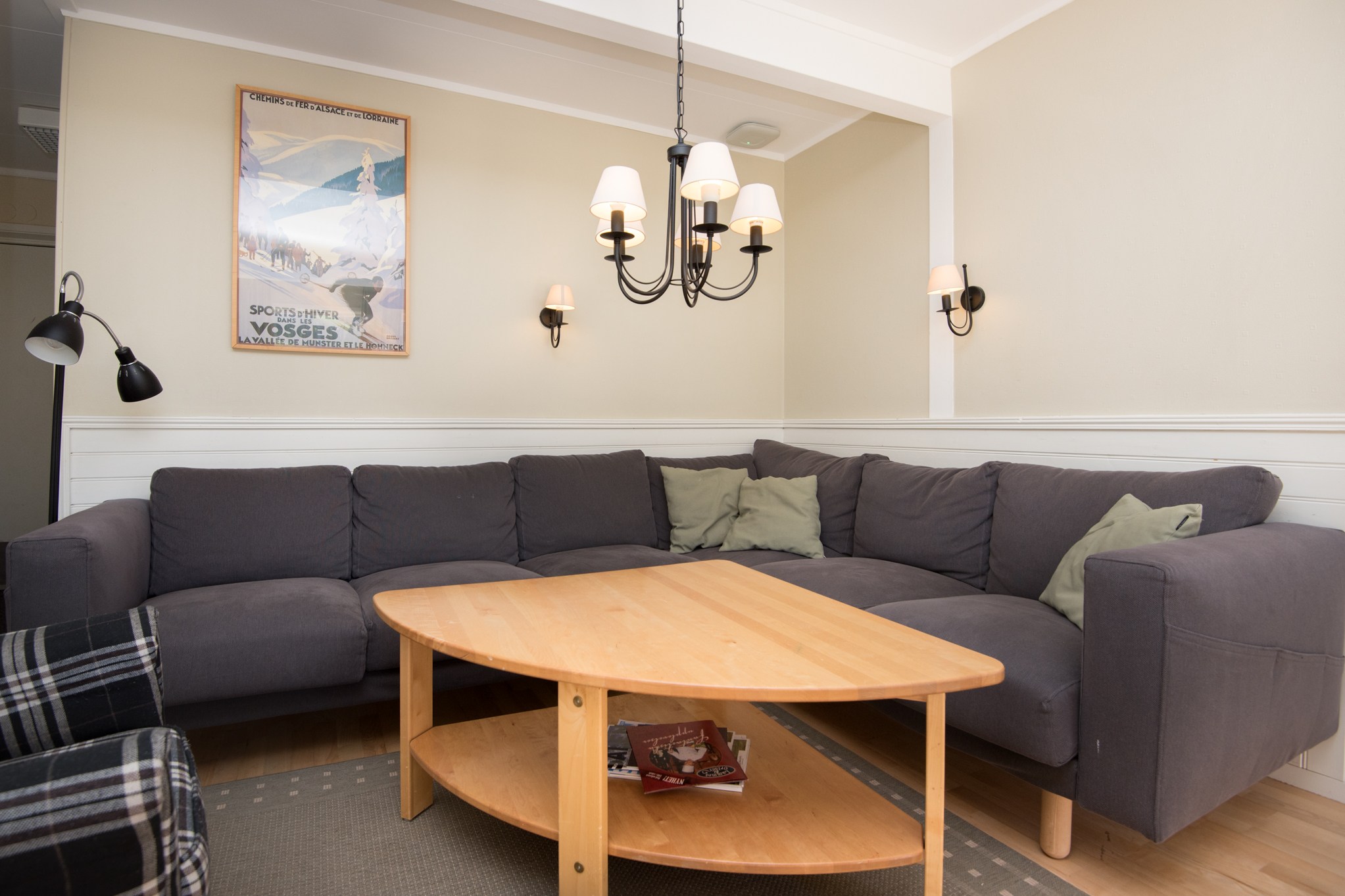En hyggelig stue med en grå L-formet sofa prydet med grønne hynder, der minder om en afslappende sommerferie i Østrig, har et lyst sofabord i træ, en plaid lænestol og en vintage plakat på væggen. En sort lysekrone hænger fra loftet og tilføjer et elegant touch.