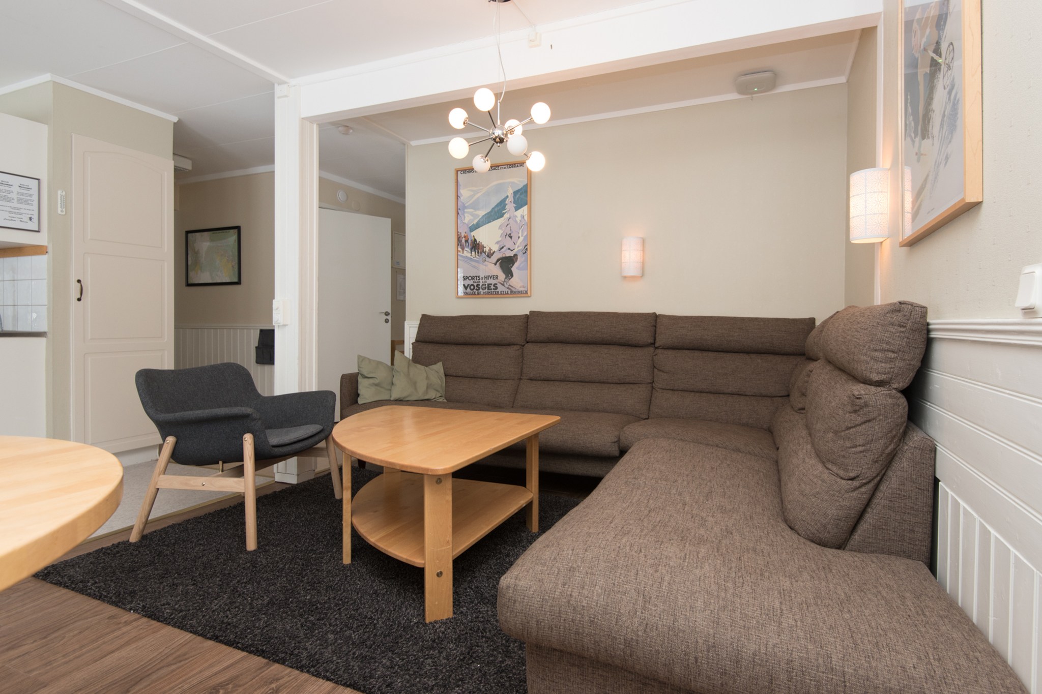 En hyggelig stue med en brun L-formet sofa og træ sofabord sætter scenen, der minder om afterski afslapning. En sort lænestol på et matchende tæppe tilføjer charme, mens moderne lysarmaturer oplyser indrammede kunstværker, der minder om Wagrains maleriske landskaber mod de beige vægge.