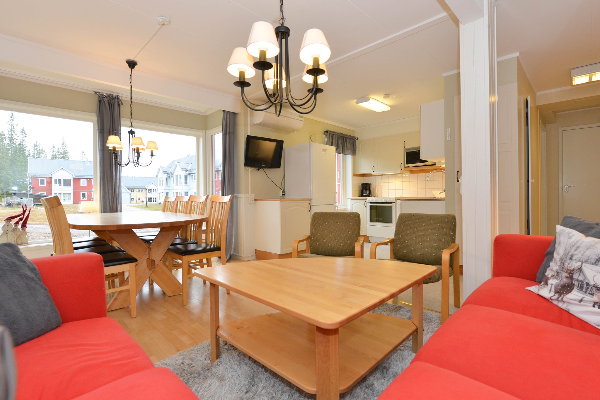 En hyggelig stue og køkkenafdeling perfekt til sommerferie Østrig, med røde sofaer, et sofabord i træ og et spisebord ved store vinduespartier. Køkkenet har hvide skabe og hårde hvidevarer. En lysekrone pryder loftet med udsigt over små huse, der minder om charmerende Wagrain.