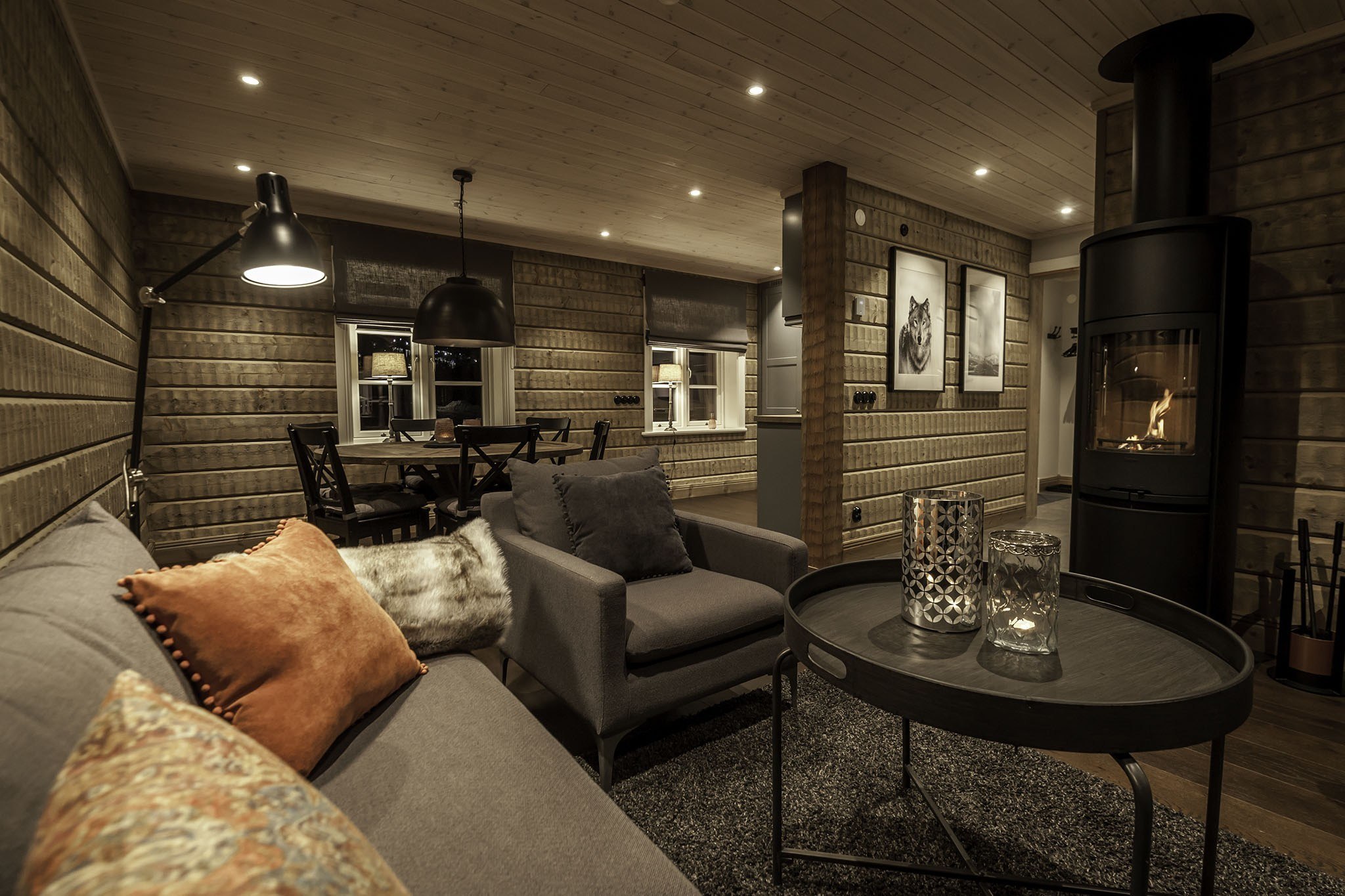 En hyggelig, varmt oplyst stue med trævægge fremkalder charmen ved Saalbach. Den moderne grå sofa og lænestol er fremhævet med hynder, mens et rundt sofabord rummer dekorative genstande. I baggrunden sidder et spisebord under en hængende lampe ved siden af en subtilt glødende pejs.