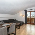 Hyggelig loftsstue med sort sektionssofa, sofabord i træ og spisebord. Et skrå loft og vægmonterede lamper tilføjer karakter. Store glasdøre åbner ud til en altan, med udsigt til sneklædte bjerge perfekt til planlægning af din næste skiferie. Lyse trægulve overalt.