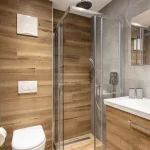Moderne badeværelse, der minder om en luksuriøs skiferiehytte, med træ og grå flisevægge. Det inkluderer brusekabine af glas med regnbrusehoved, hvidt toilet og trævask med hvid vask. Det overordnede design er slankt og minimalistisk, hvilket fremkalder elegancen ved Wagrains alpine retreater.