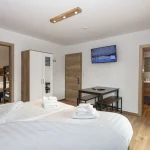 Et moderne hotelværelse i Westendorf med en hyggelig dobbeltseng dækket af hvidt sengetøj og håndklæder. Et lille bord med to bænke støder op, perfekt til planlægning af dine sommerferie Østrig eventyr. Et fladskærms-tv er monteret på væggen over bordet. Værelset har også et klædeskab og trædetaljer.